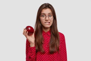 Mulher jovem com óculos e expressão de dúvida segurando uma maçã vermelha, ilustrando restrições alimentares no clareamento dental na Cuidalli Odontologia, no bairro da Saúde