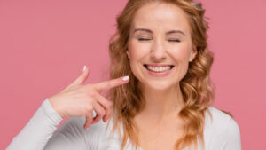 Mulher loira sorrindo com olhos fechados aponta para os dentes. Implante dentário unitário na Cuidalli Saúde para evitar dentes tortos.