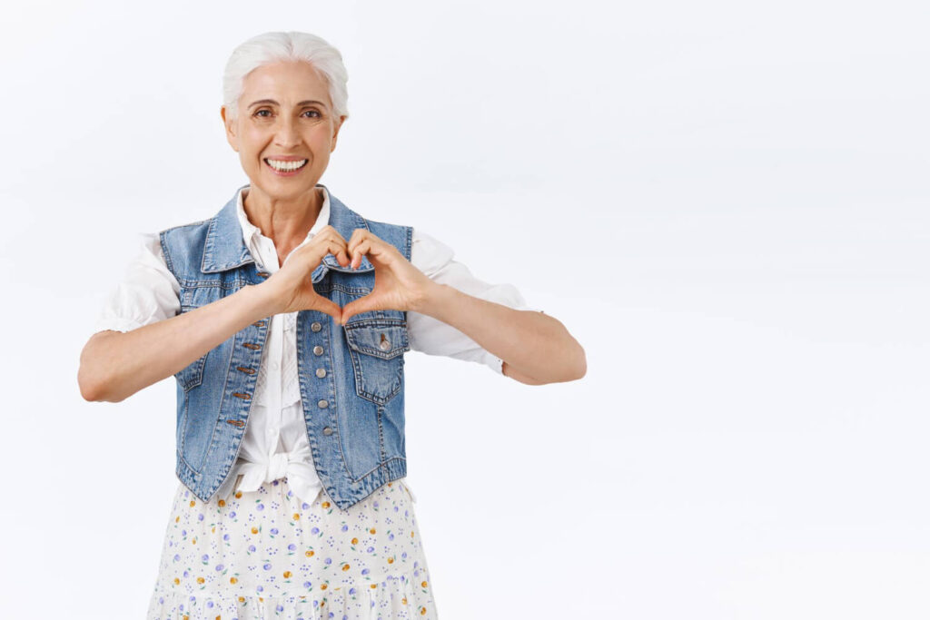 Mulher idosa sorridente com cabelos brancos faz um gesto de coração com as mãos. Cuidalli Odontologia: saúde bucal e prevenção cardíaca.