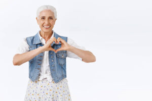 Mulher idosa sorridente com cabelos brancos faz um gesto de coração com as mãos. Cuidalli Odontologia: saúde bucal e prevenção cardíaca.
