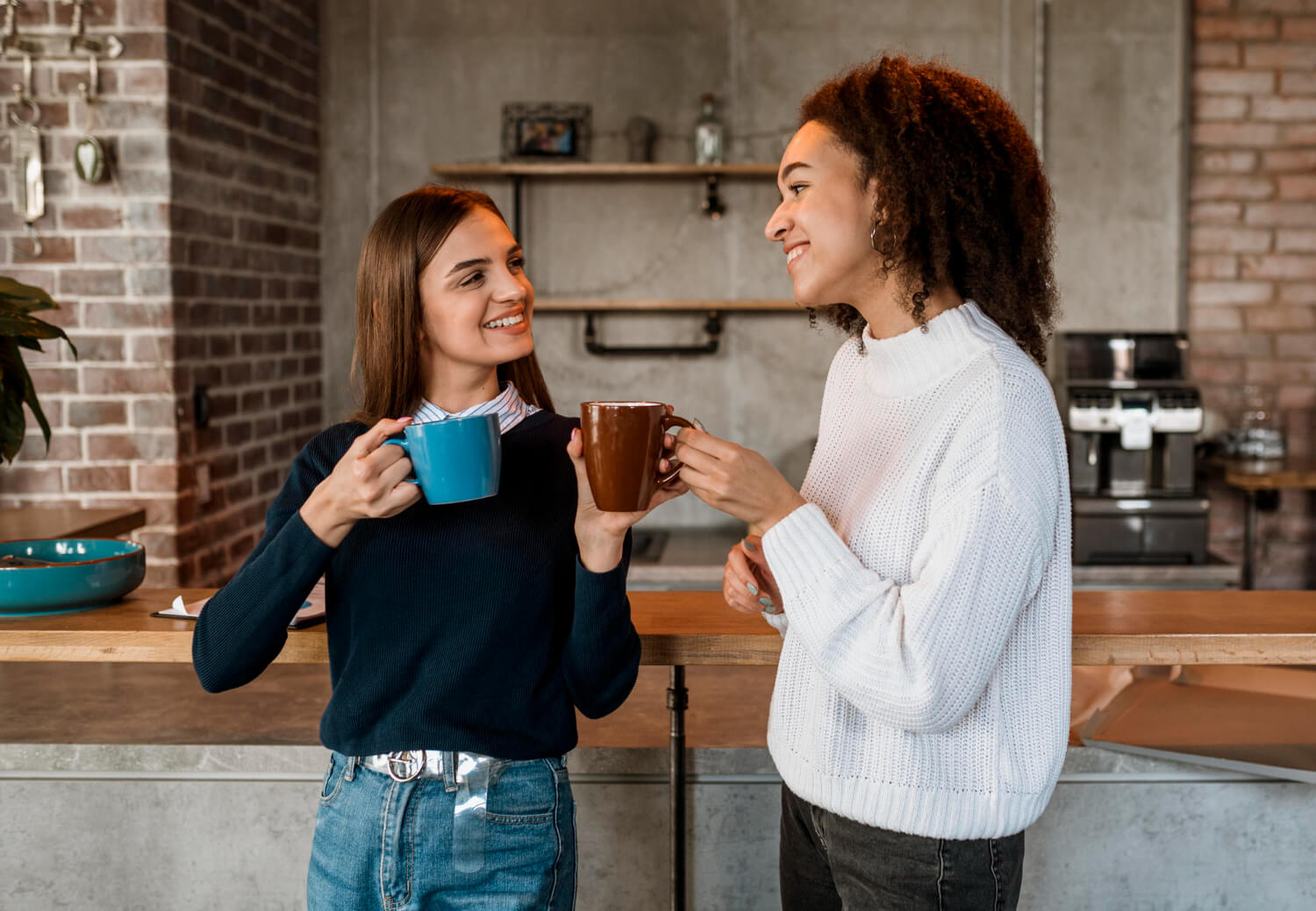 Duas mulheres sorridentes segurando canecas de café em uma cozinha moderna, discutindo sobre a saúde e manchas em lentes de contato dental na Clínica Cuidalli.