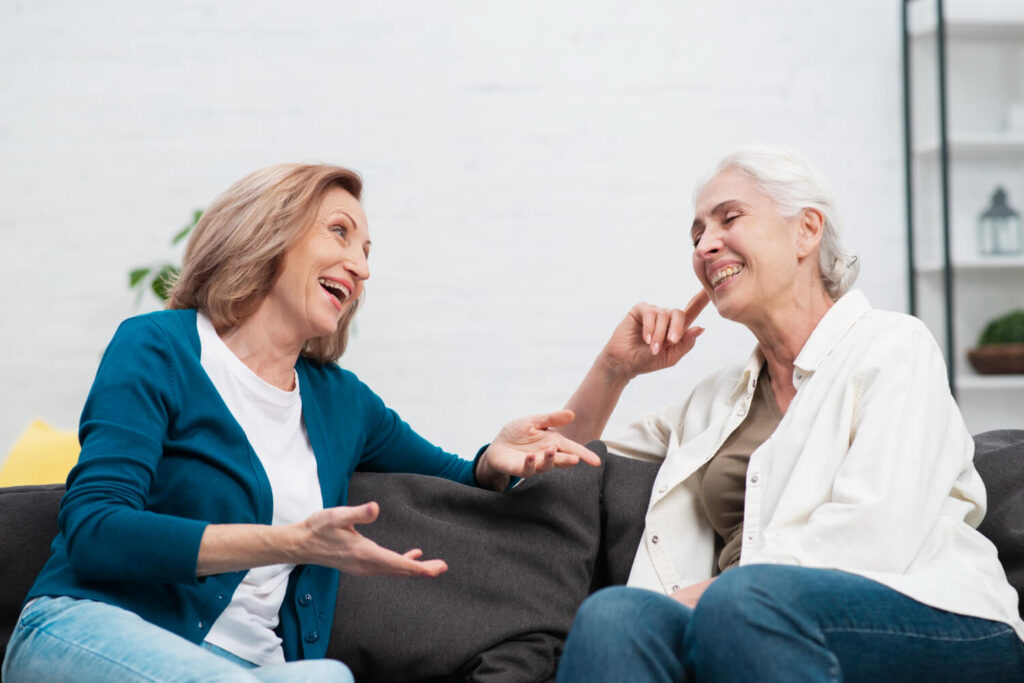 Duas idosas conversam e riem sentadas no sofá, mostrando a segurança de quem usa prótese protocolo em vez de dentadura comum na Saúde.