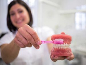 Dentista sorrindo demonstra como higienizar corretamente a prótese protocolo usando uma escova rosa em modelo dentário na Saúde, SP.